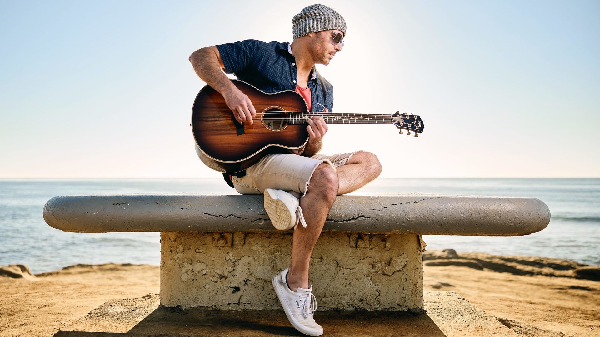 man playing Taylor GT K21e guitar on cement pedestal at the beach