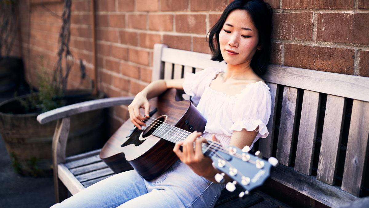 woman playing Taylor AD27 acoustic guitar on bench