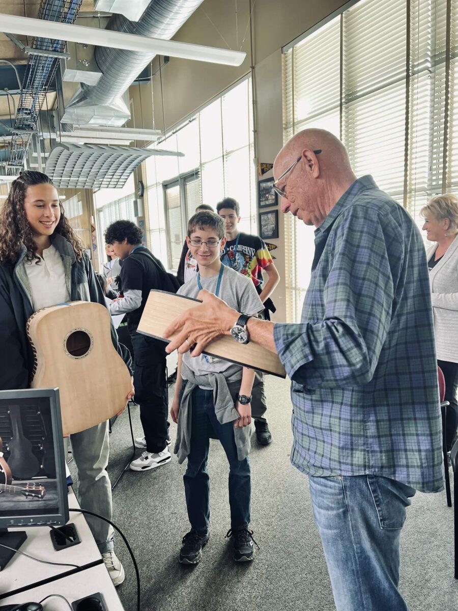 Bob Taylor holds body of acoustic guitar while talking to smiling high school wood shop students